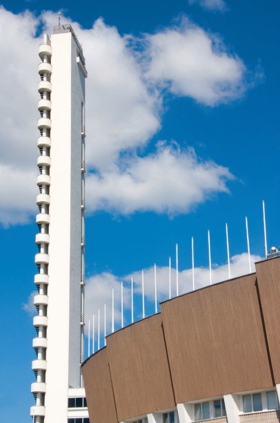 Kõrge huvitava arhitektuuriga valge torn madalama pruuni staadioniääre kõrval, tagaplaanil mõne pilvegasinine taevas