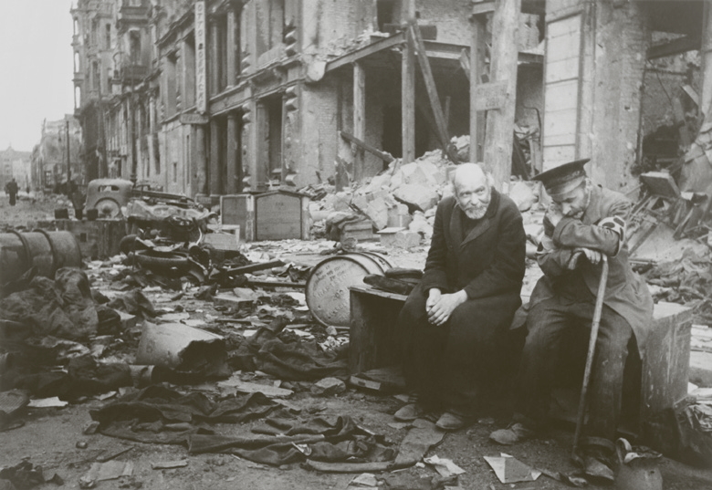 Vana mees istumas purustuste keskel pingil, tema ümber palju sodi ja seljataga maja varemed